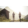 Zwei Menschen joggen in der Abendsonne einen Weg entlang, im Hintergrund ein Bergmassiv und eine Hütte / Two people jog along a path in the evening sun, with a mountain massif and a hut in the background