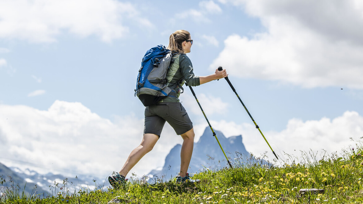 Ein Mann mit Rucksack und Wanderstöcken steigt über eine Wiese bergauf, im Hintergrund ein felsiger Gipfel / A man with a backpack and trekking poles hikes uphill across a meadow with a rocky peak in the background
