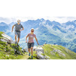 Zwei Männer laufen bei Tageslicht auf einem Bergpfad mit Aussicht auf grüne Alpengipfel / Two men run on a mountain trail with views of green alpine peaks