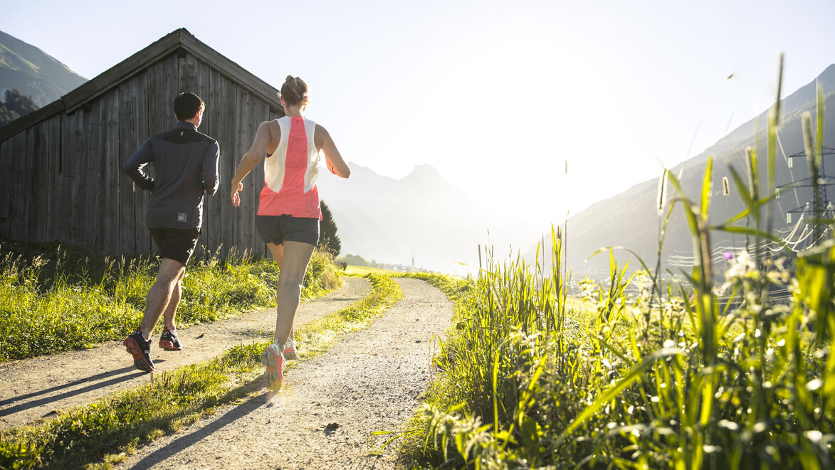 Zwei Personen laufen auf einem sonnigen Feldweg an einer Holzhütte vorbei in Richtung Berge / Two people run on a sunny field path past a wooden hut towards the mountains