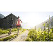Zwei Personen laufen auf einem sonnigen Feldweg an einer Holzhütte vorbei in Richtung Berge / Two people run on a sunny field path past a wooden hut towards the mountains