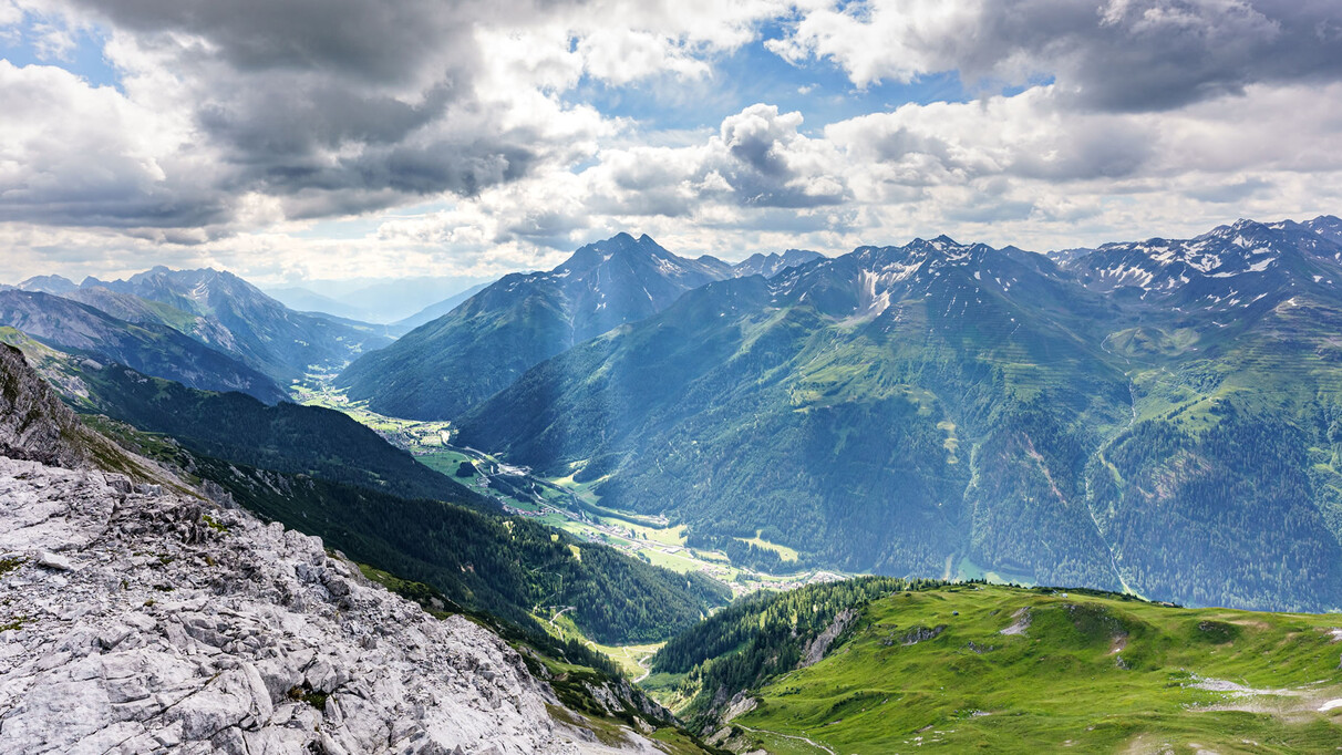 Weitwinkelaufnahme von St. Anton am Arlberg mit Blick über das Stanzertal, umrahmt von bewaldeten Bergen und Sonnenstrahlen zwischen den Wolken.