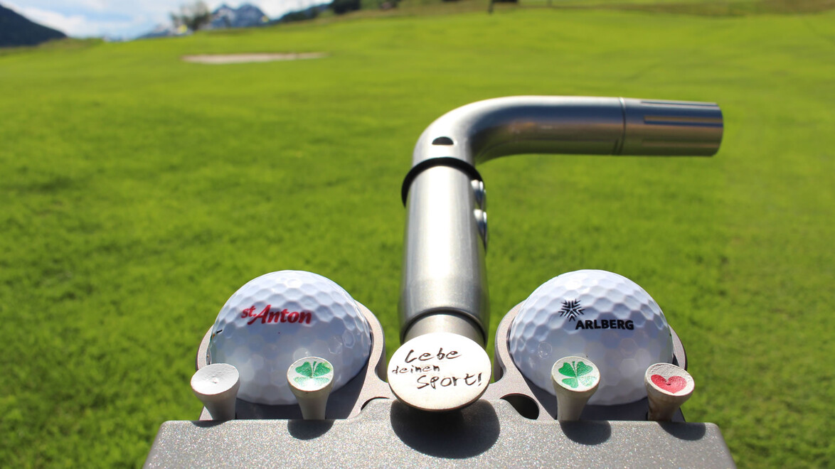 Zwei Golfbälle mit Abschlagsmarkierungen liegen auf einer Ballausgabestation am Golfplatz. / Two golf balls with tee markers lie on a ball dispenser at the golf course.