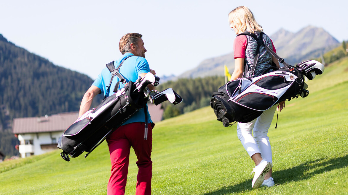 Ein Mann und eine Frau tragen ihre Golfschläger-Taschen über den Golfplatz mit Blick auf Berge. / A man and a woman carry their golf bags across the course with mountain views.