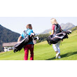 Ein Mann und eine Frau tragen ihre Golfschläger-Taschen über den Golfplatz mit Blick auf Berge. / A man and a woman carry their golf bags across the course with mountain views.