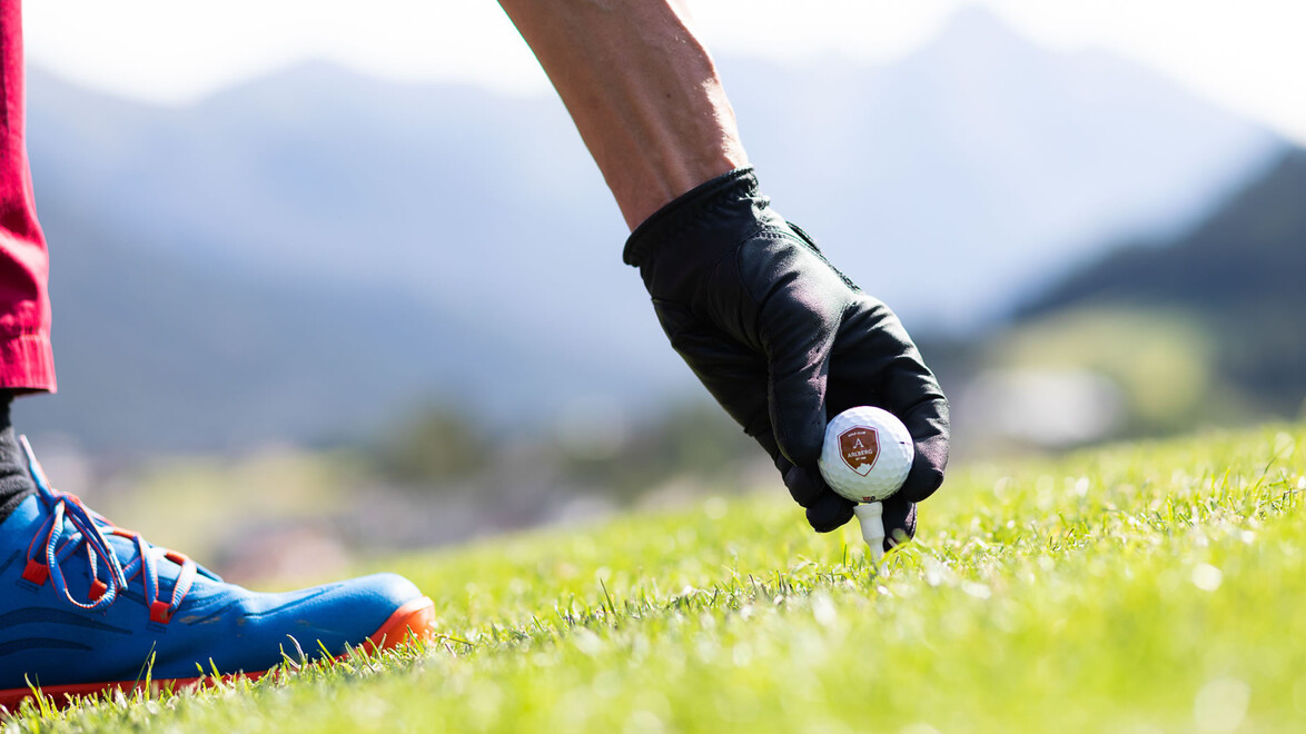 Eine Hand mit Golfhandschuh platziert einen Golfball auf einem Tee im Gras. / A gloved hand places a golf ball on a tee on the grass.