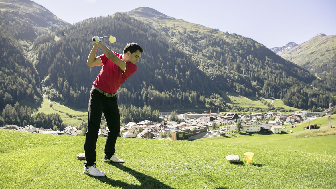 Ein Golfer schlägt am Abschlag mit Blick auf ein Bergdorf im Hintergrund. / A golfer tees off with a mountain village in the background.