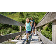 Ein Mädchen läuft über eine Holzbrücke, gefolgt von einem Erwachsenen in sommerlicher Berglandschaft / A girl runs across a wooden bridge followed by an adult in a summer mountain setting