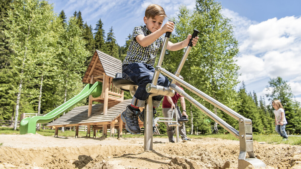 Ein Kind spielt auf einem Baggergerät im Sandspielbereich vor einem Holzspielplatz mit Rutsche / A child operates a digger toy in a sandy play area in front of a wooden playground with slide