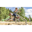 Ein Kind spielt auf einem Baggergerät im Sandspielbereich vor einem Holzspielplatz mit Rutsche / A child operates a digger toy in a sandy play area in front of a wooden playground with slide