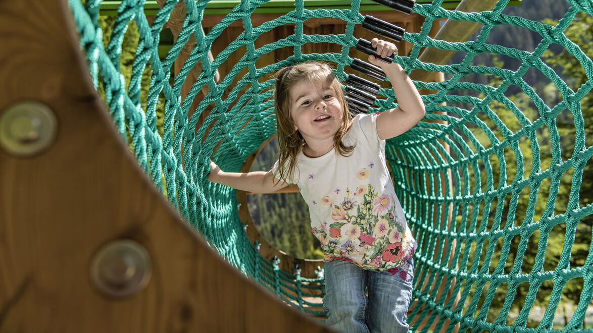 Ein Mädchen krabbelt lachend durch ein grünes Kletternetz an einem Spielplatz / A girl crawls smiling through a green climbing net at a playground