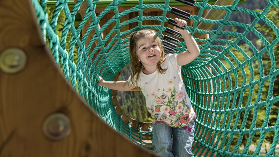 A girl smiles while crawling through a green rope tunnel on a playground, with alpine views in the background.