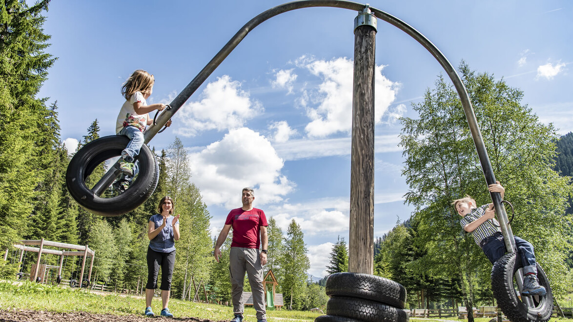 Zwei Kinder schwingen auf Reifenschaukeln, während Erwachsene daneben stehen, auf einer Wiese im Wald / Two children swing on tire swings while adults stand nearby on a forest meadow