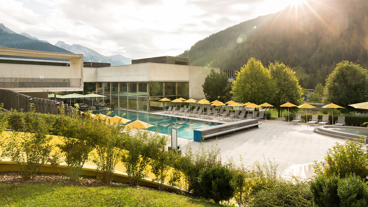 The outdoor pool of Arlberg WellCom in St. Anton am Arlberg, with yellow umbrellas, modern buildings and alpine greenery.