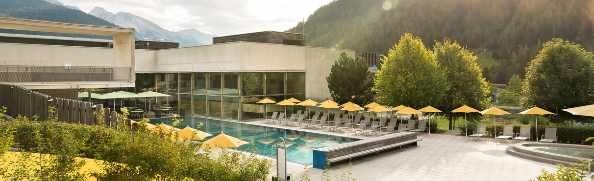 The outdoor pool of Arlberg WellCom in St. Anton am Arlberg, with yellow umbrellas, modern buildings and alpine greenery.