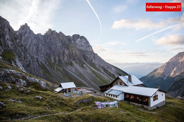Eine Alpenhütte liegt unterhalb steiler Felswände mit weiter Aussicht ins Tal bei Morgendämmerung / An alpine hut sits below steep rock faces with a wide valley view at dawn