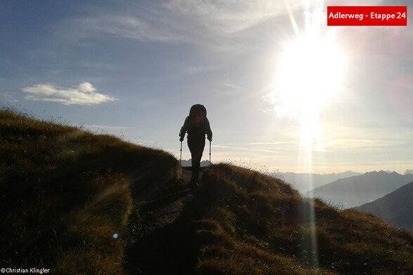 Wanderer im Gegenlicht auf einem grasbewachsenen Bergpfad mit Sonne am Horizont / Hiker in backlight on a grassy mountain trail with the sun on the horizon