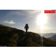 Wanderer im Gegenlicht auf einem grasbewachsenen Bergpfad mit Sonne am Horizont / Hiker in backlight on a grassy mountain trail with the sun on the horizon