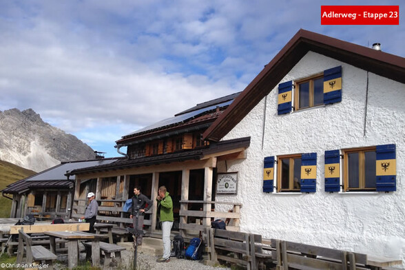 Eine Alpenhütte mit Terrasse und Wanderern davor, im Hintergrund felsige Bergkulisse / An alpine hut with a terrace and hikers in front, rocky mountains in the background