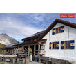 Eine Alpenhütte mit Terrasse und Wanderern davor, im Hintergrund felsige Bergkulisse / An alpine hut with a terrace and hikers in front, rocky mountains in the background