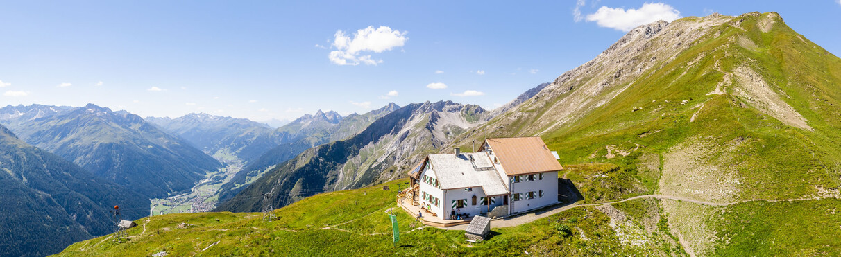 Weiße Ansbacher Hütte mit braunem Dach auf grünem Berghang, im Hintergrund hohe Berge und ein Tal.