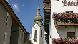 Der barocke Kirchturm von Flirsch ragt zwischen Häusern mit Blumenbalkonen in den Himmel.
