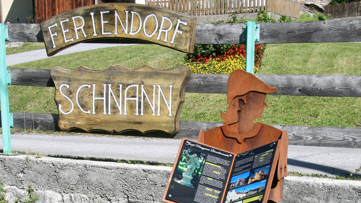 Themenweg-Figur mit Buch steht vor einem Holzschild mit „Feriendorf Schnann“ / Themed trail figure with book in front of a wooden sign reading “Feriendorf Schnann”