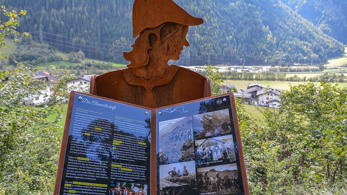 Rostfarbene Figur mit bebildertem Buch über Schnann im Vordergrund, Dorf im Tal dahinter / Rust-colored figure with illustrated book about Schnann in foreground, village in the valley behind