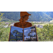 Rostfarbene Figur mit bebildertem Buch über Schnann im Vordergrund, Dorf im Tal dahinter / Rust-colored figure with illustrated book about Schnann in foreground, village in the valley behind