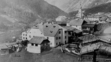 Ein schwarz-weißes Archivfoto zeigt das Dorf Flirsch bei St. Anton am Arlberg im 19. Jahrhundert mit traditionellen Steinhäusern und schindelgedeckten Dächern.