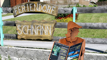 Eine Holztafel begrüßt Besucher im Feriendorf Schnann bei St. Anton am Arlberg. Ein stilisierter Detektiv weist auf den Themenweg durch das Dorf hin.