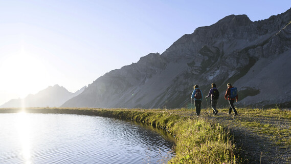 Drei Wanderer gehen am Ufer eines ruhigen Speichersees bei St. Anton am Arlberg entlang, während die Sonne über die Berggipfel steigt.