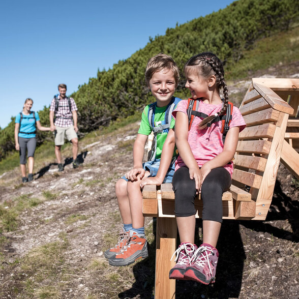 Zwei Kinder sitzen lachend auf einer hölzernen Panoramabank am Wanderweg oberhalb von St. Anton am Arlberg, während ihre Eltern im Hintergrund den Hang heraufkommen. Sonniger Tag inmitten grüner Alpenlandschaft.