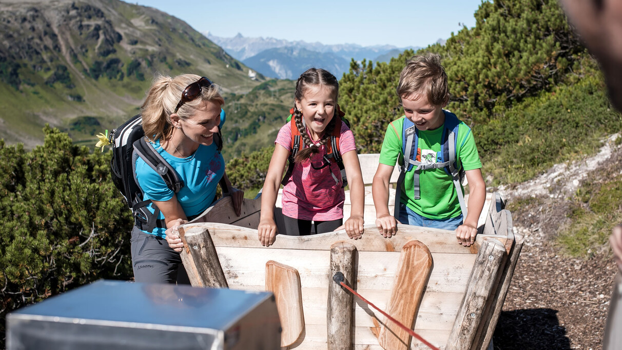Two backpacked children and their mother laugh while exploring a wooden play station during a hike above St. Anton am Arlberg. In the background: green alpine slopes and clear skies.