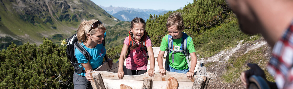 Zwei Kinder sitzen lachend auf einer hölzernen Panoramabank am Wanderweg oberhalb von St. Anton am Arlberg, während ihre Eltern im Hintergrund den Hang heraufkommen. Sonniger Tag inmitten grüner Alpenlandschaft.