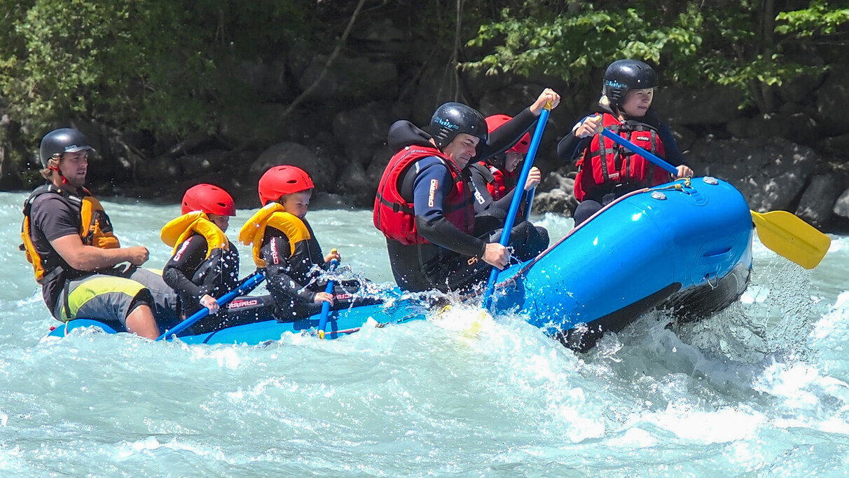 Familie mit Kindern beim Rafting auf der Rosanna bei St. Anton am Arlberg. Im blauen Boot paddeln alle mit Helmen und Schwimmwesten durch das spritzende Wildwasser.