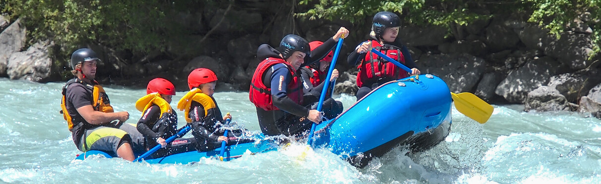 Familie mit Kindern beim Rafting auf der Rosanna bei St. Anton am Arlberg. Im blauen Boot paddeln alle mit Helmen und Schwimmwesten durch das spritzende Wildwasser.