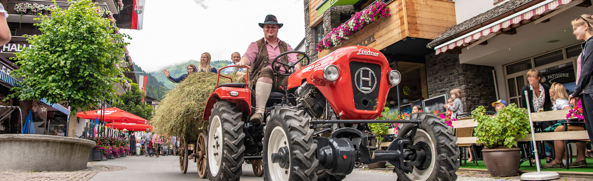 Ein Mann in traditioneller Tiroler Kleidung fährt mit einem roten Oldtimer-Traktor samt Heuladung durch die Fußgängerzone von St. Anton.