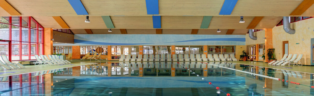 Das Hallenbad im Arlberg WellCom in St. Anton am Arlberg mit Schwimmbahnen, vielen Liegen, Glasfassade und farbigen Deckenelementen.