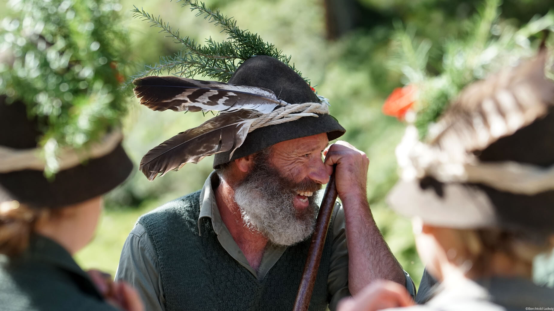 Ein älterer Mann mit traditionellem Hut und Hahnenfedern lacht im Gespräch bei einer Festveranstaltung. / An older man with a traditional hat and rooster feathers laughs in conversation at a festive event.