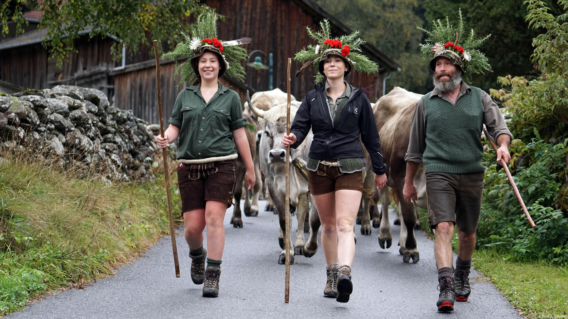 Drei Personen in Tracht stehen mit Hirtenstöcken am Wegrand, bereit für den Almabtrieb. / Three people in traditional clothing stand with herding sticks at the roadside, ready for the cattle drive.
