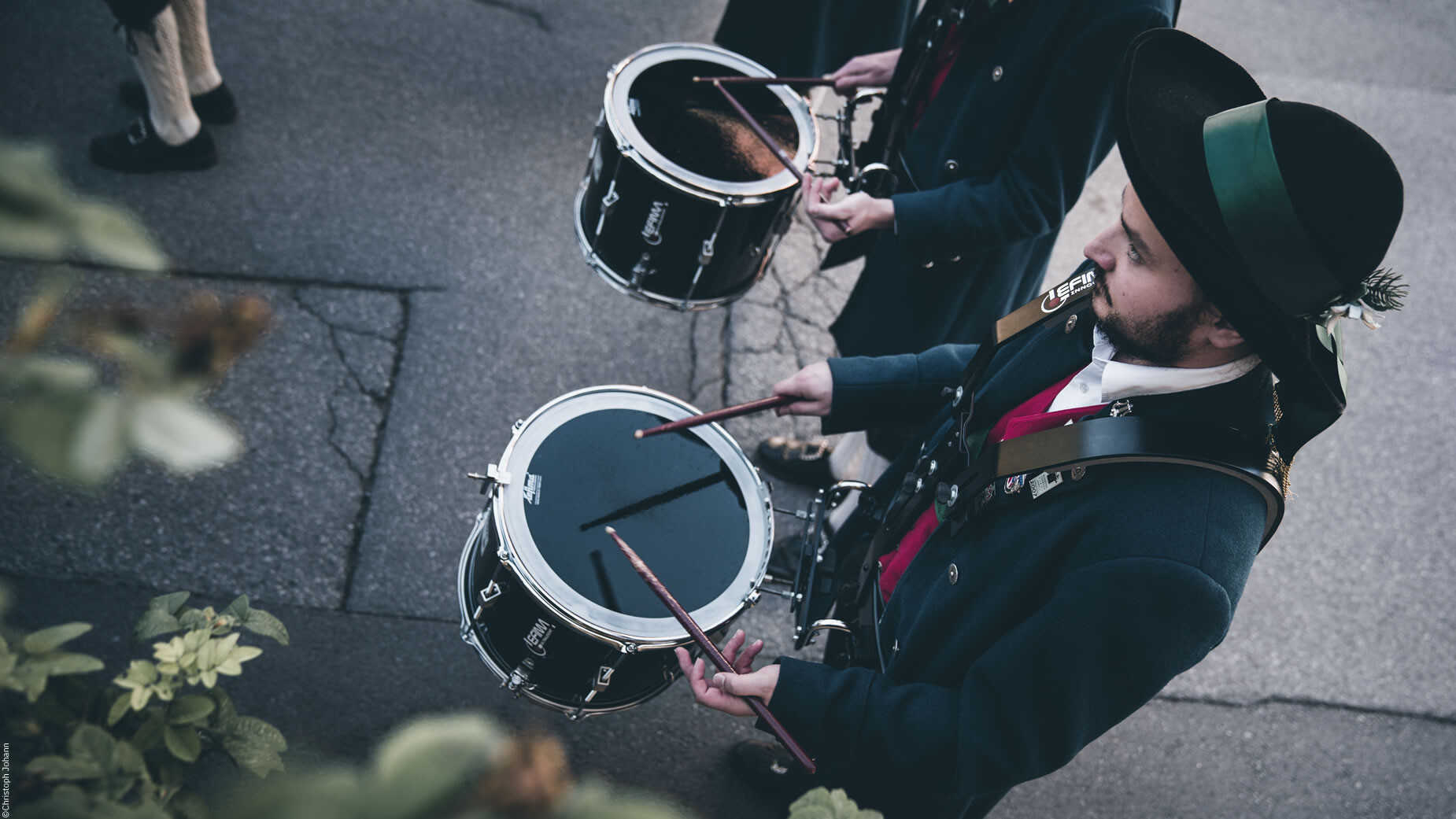 Zwei Musiker in Tracht spielen während des Umzugs auf kleinen Marschtrommeln. / Two musicians in traditional attire play marching drums during the parade.