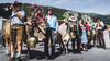 Mehrere festlich geschmückte Kühe stehen mit ihren Begleitpersonen vor einer Bergkulisse. / Several festively adorned cows stand with their handlers in front of a mountain backdrop.