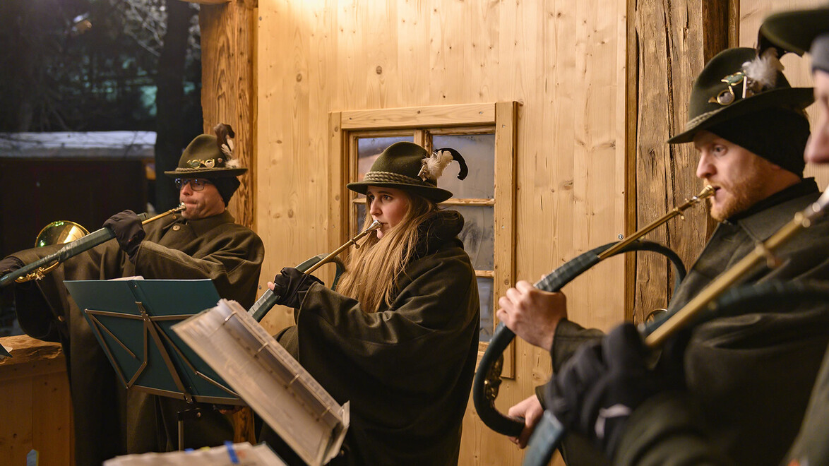 Bläsergruppe mit traditionellen Hüten spielt in weihnachtlich beleuchtetem Holzstand / Brass band in traditional hats plays in a Christmas-lit wooden stall