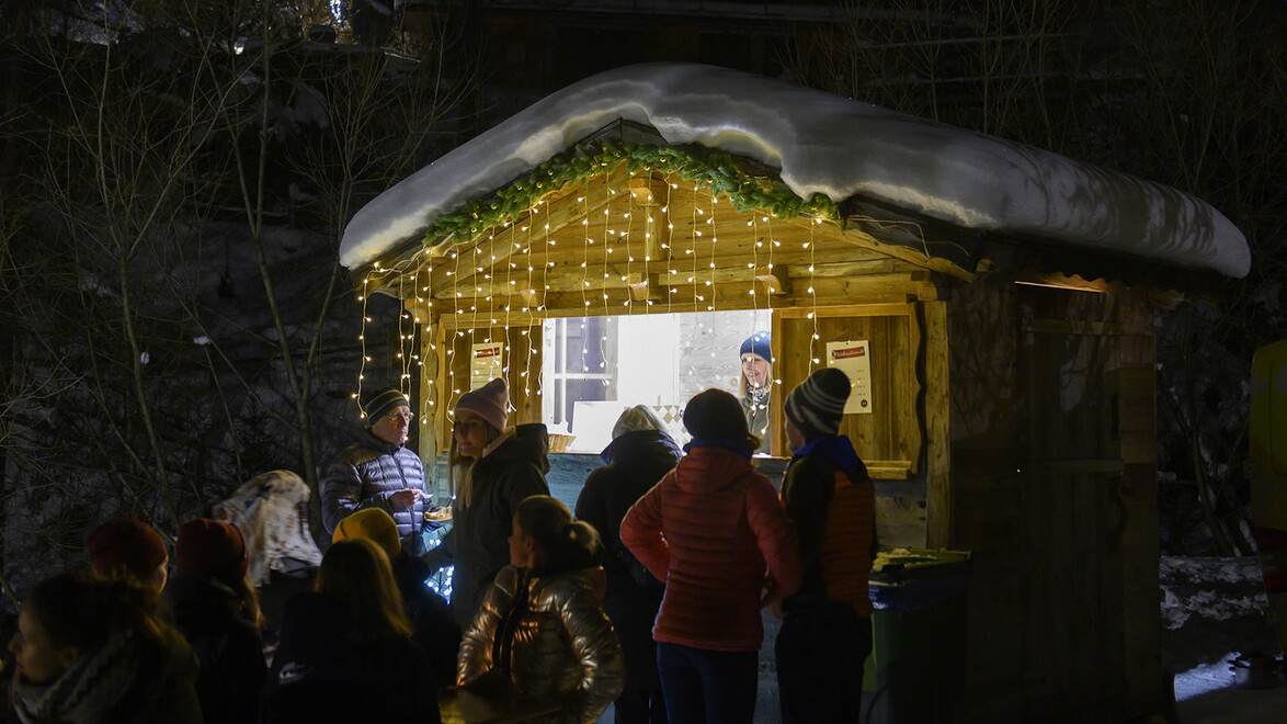 Besucher:innen stehen an beleuchtetem Verkaufsstand mit Lichterkette im Schnee / Visitors at illuminated market stall with fairy lights in the snow