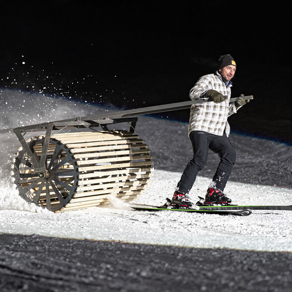 Mann auf Skiern zieht eine hölzerne Pistenwalze über die Schneepiste.