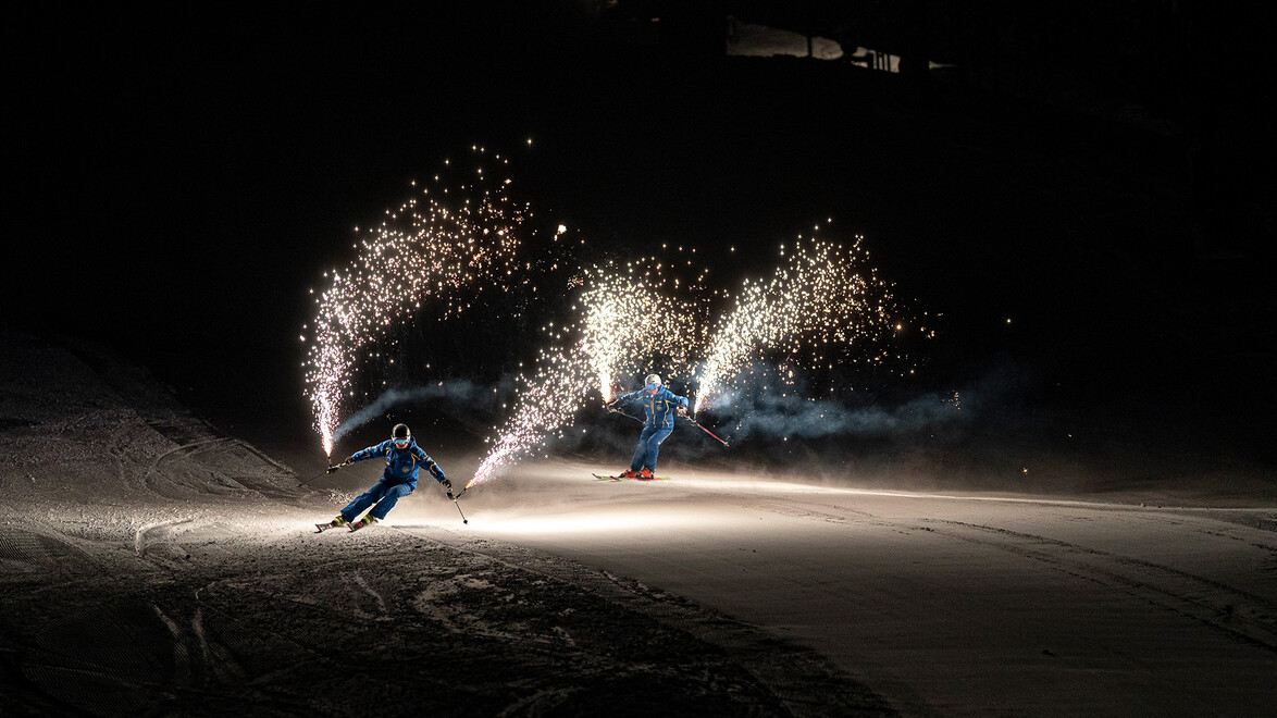 Drei Skifahrer:innen ziehen bei Nacht mit Pyroeffekten leuchtende Spuren im Schnee. / Three skiers create glowing tracks with pyrotechnics at night.