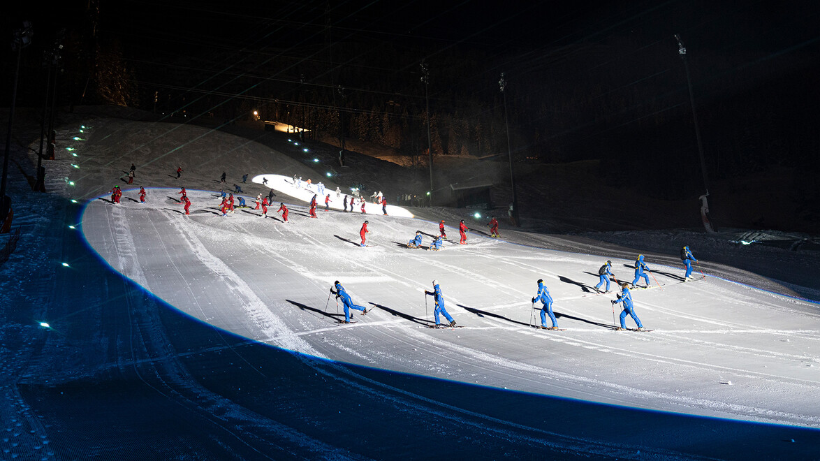 Ski-Instruktor:innen in blauer Kleidung zeigen eine Formation auf der nächtlich beleuchteten Skipiste. / Ski instructors in blue outfits perform a formation on a floodlit slope.