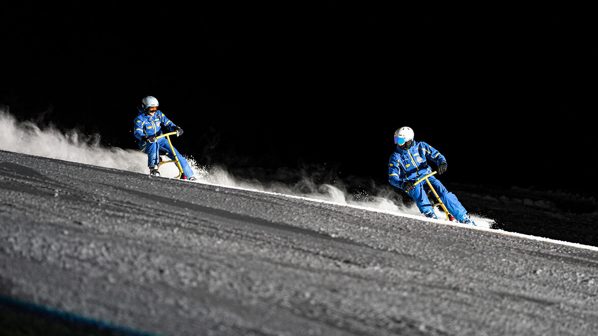 Zwei Skifahrer:innen auf Snowbikes fahren bei Nacht rasant die Piste hinunter. / Two snowbikers race downhill on a night slope.