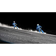 Zwei Skifahrer:innen auf Snowbikes fahren bei Nacht rasant die Piste hinunter. / Two snowbikers race downhill on a night slope.